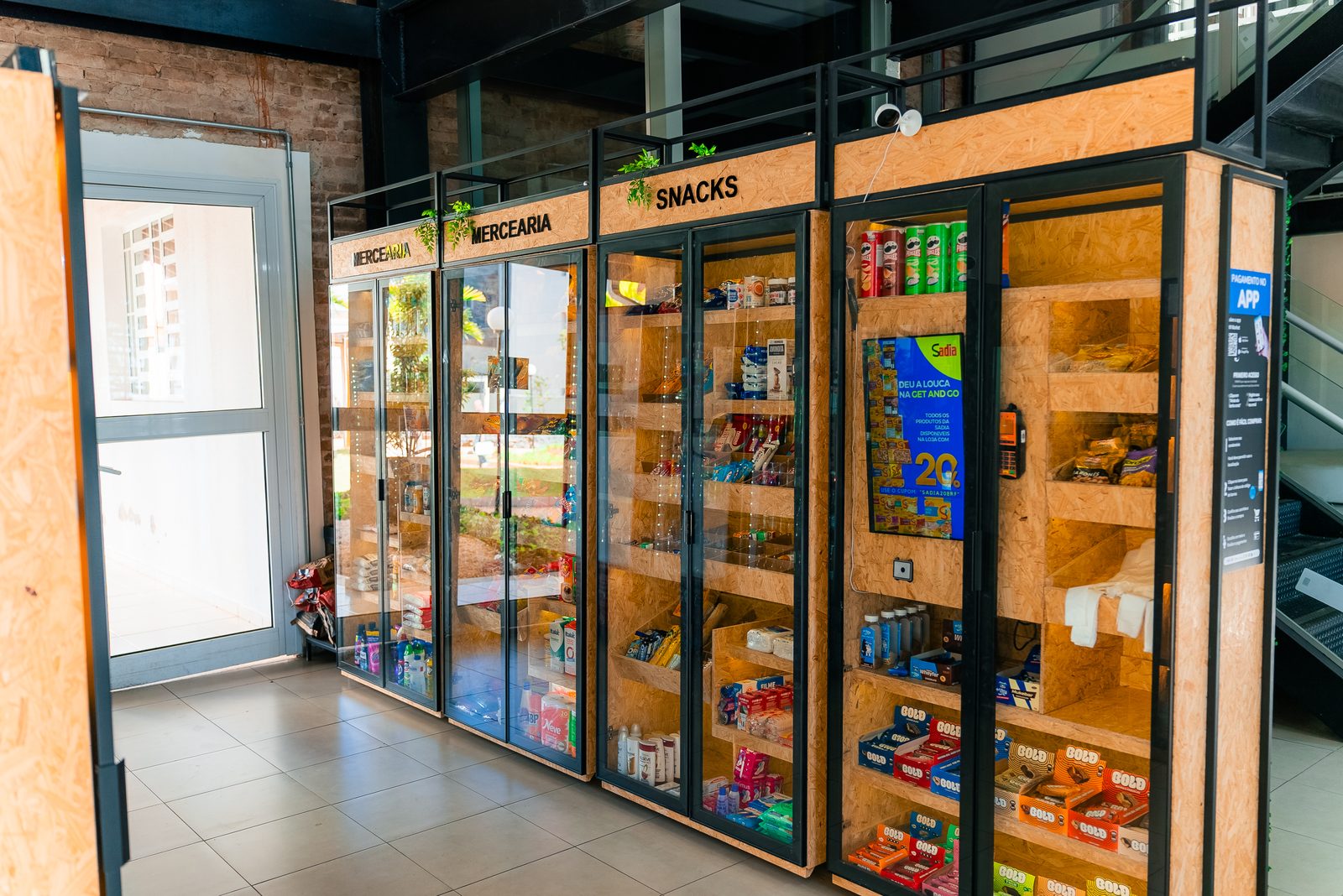 Vitrines de vidro e madeira de uma loja autônoma, com prateleiras de mercearia e snacks iluminadas