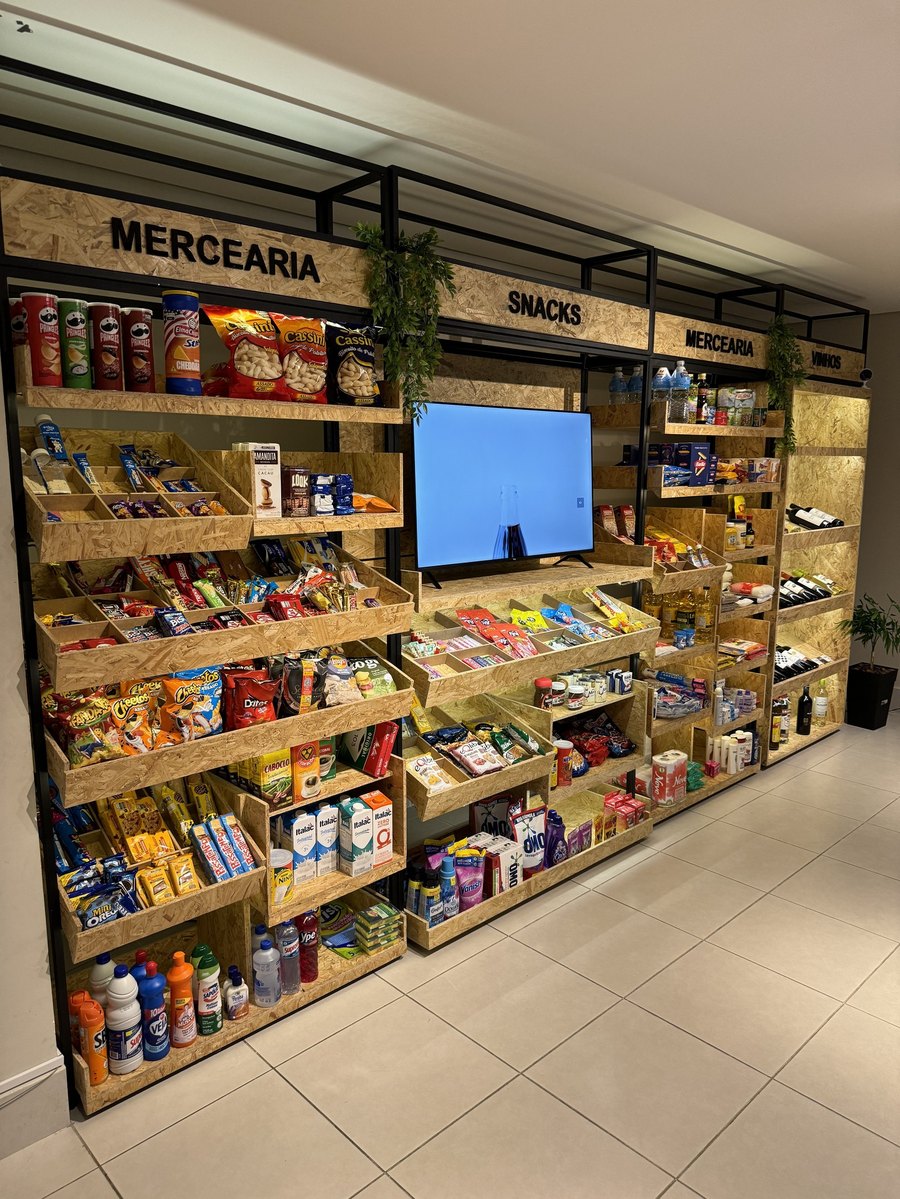 Parede de gôndolas de madeira com mercearia, snacks e vinhos, sinalizada por categoria